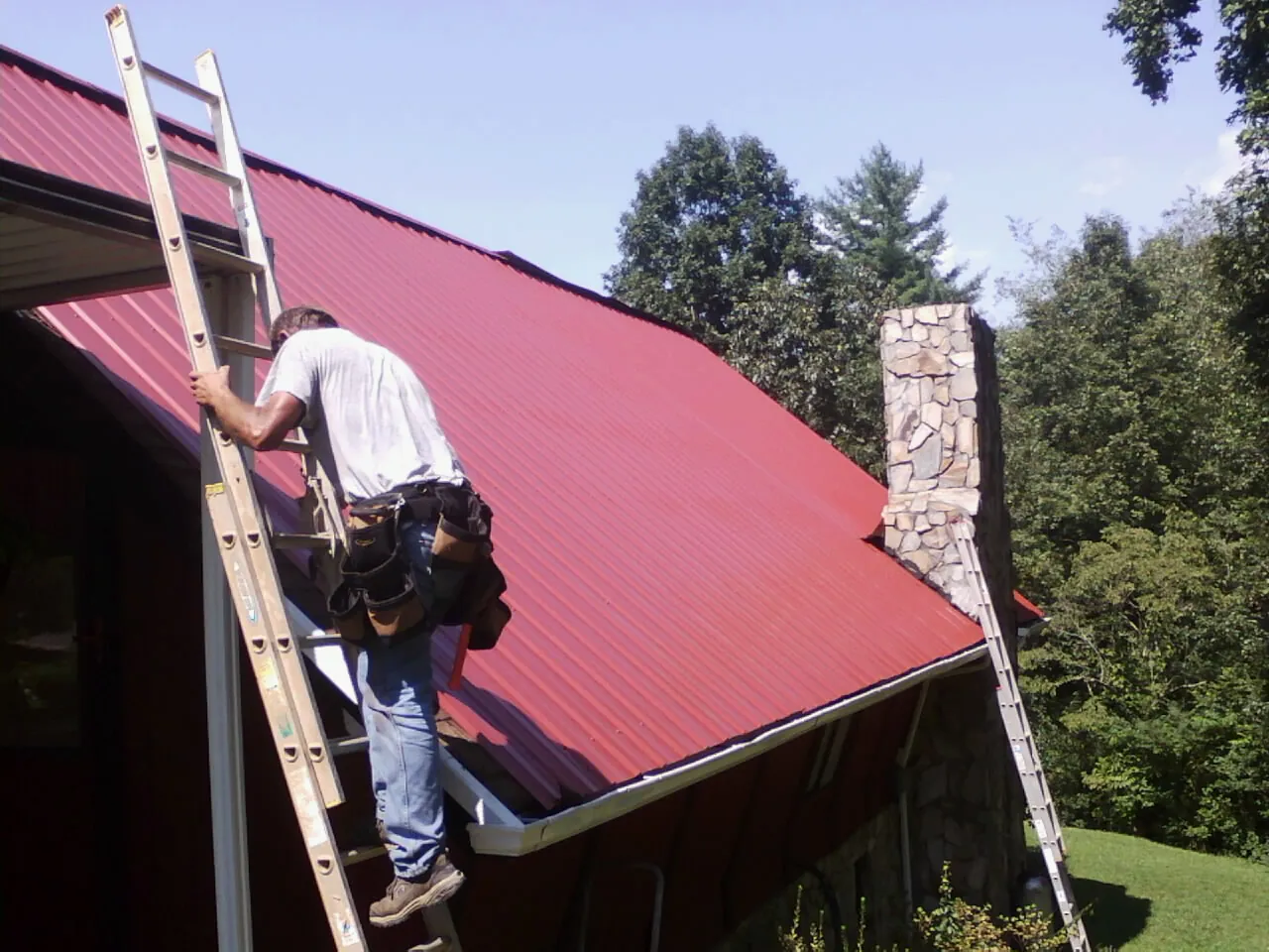 Roofer on a ladder beside a red metal roof and stone chimney — Soffit & Fascia Repair by Roofing Brothers in Electric City